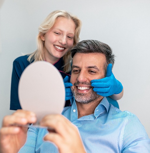 Dentist with blue gloves touching patient’s smile as he looks into mirror