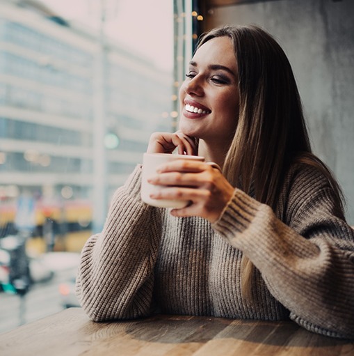 Smiling woman in brown sweater with coffee mug looking out window
